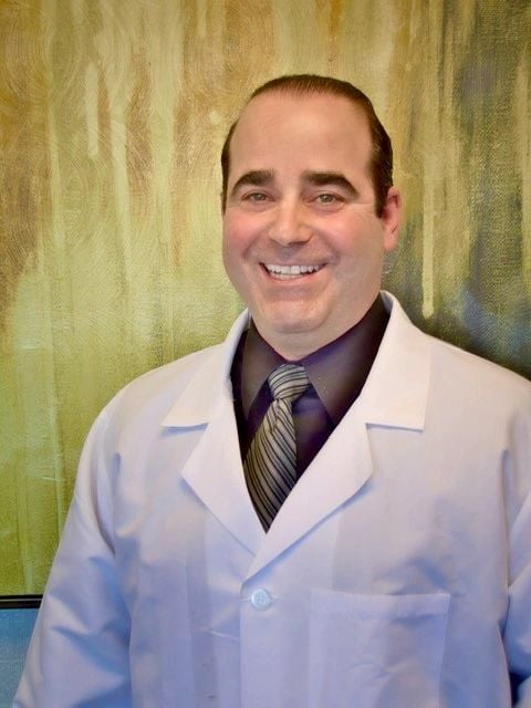 Dr. Brian Opensky, board-certified podiatrist, smiling in a white coat against a textured green background, representing Opensky Podiatry's commitment to personalized foot care.