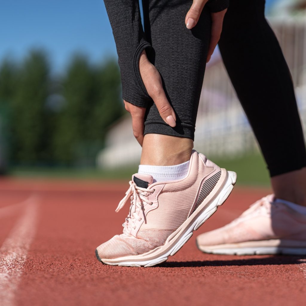 Woman in athletic wear touching her ankle in pain on a running track, illustrating an ankle sprain related to sports injuries, relevant to podiatric care services offered by Opensky Podiatry.